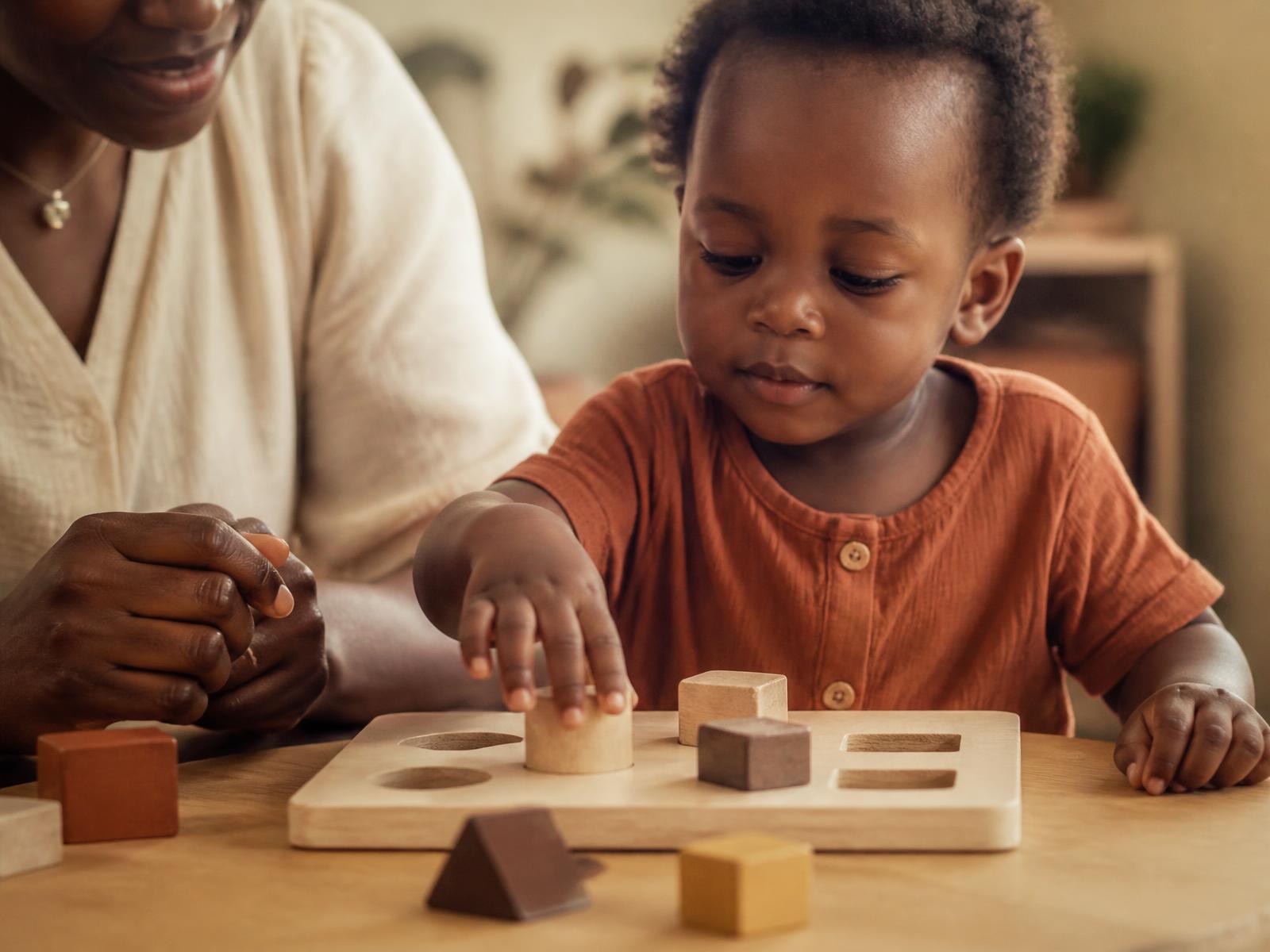 A Nigerian mother beside her toddler arranging wooden shape blocks on a table